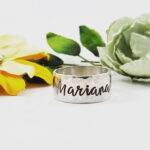 The Sterling Silver Wide Band Personalize ring, engraved with "Mariana," stands upright. Behind it are a yellow fabric flower and a pale green flower with green leaves, all set against a white background. Customize yours today!. Read more at Stamping Cat Studio (stampingcatstudio.com).