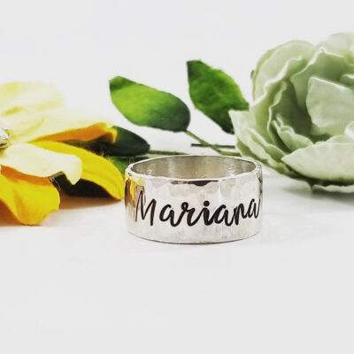 The Sterling Silver Wide Band Personalize ring, engraved with "Mariana," stands upright. Behind it are a yellow fabric flower and a pale green flower with green leaves, all set against a white background. Customize yours today!. Read more at Stamping Cat Studio (stampingcatstudio.com).