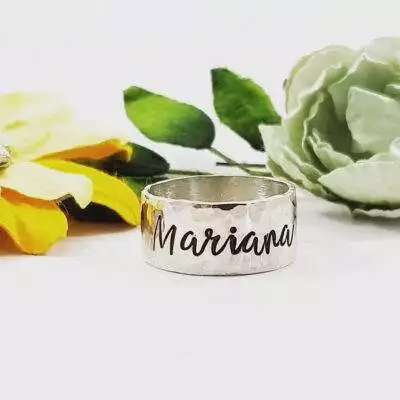 The Sterling Silver Wide Band Personalize ring, engraved with "Mariana," stands upright. Behind it are a yellow fabric flower and a pale green flower with green leaves, all set against a white background. Customize yours today!. Read more at Stamping Cat Studio (stampingcatstudio.com).