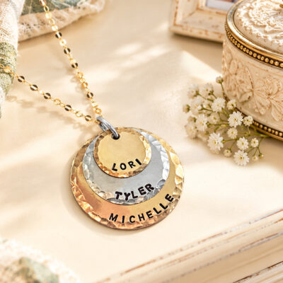 A gold chain Kids Names Neck lifestyle features three stacked, hammered metal discs engraved with the names LORI, TYLER, and MICHELLE, layered from smallest to largest against a cream-colored surface. Read more at Stamping Cat Studio (stampingcatstudio.com).