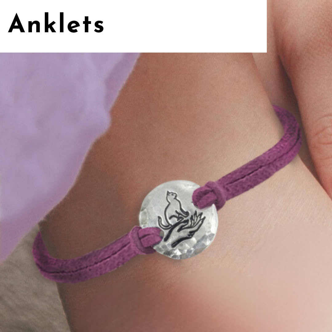 A close-up of an ankle wearing a purple anklet with a round, silver charm engraved with a howling wolf—perfect for lovers of personalized jewelry. The anklet features adjustable knots. "Anklets" appears in bold black letters on a white background, top left corner. Read more at Stamping Cat Studio (stampingcatstudio.com).