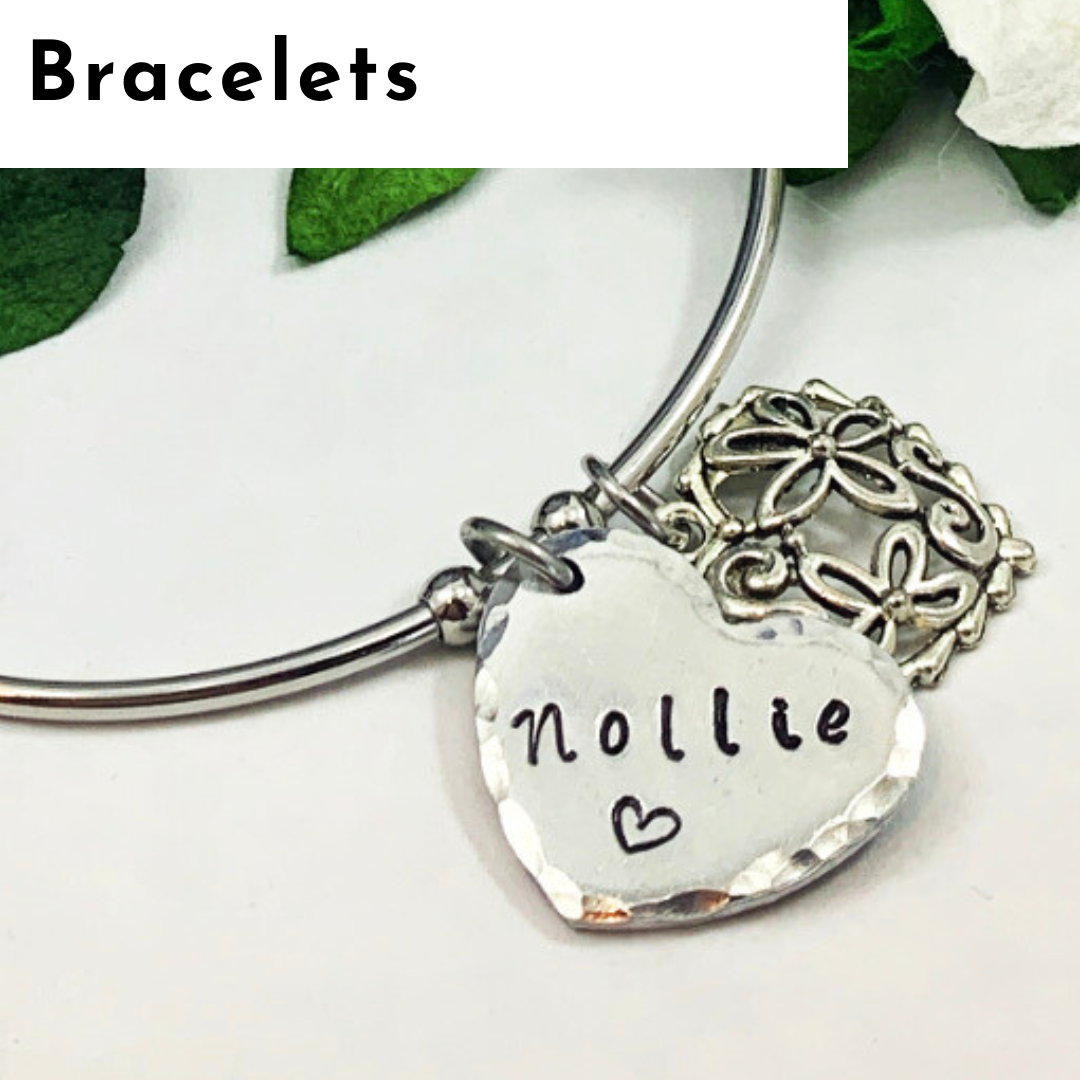 A silver bangle bracelet with two charms: a textured heart engraved with “nollie” and a small heart, plus a decorative floral charm. Green leaves and white petals set the backdrop, highlighting this piece of personalized jewelry. “Bracelets” appears at the top. Read more at Stamping Cat Studio (stampingcatstudio.com).