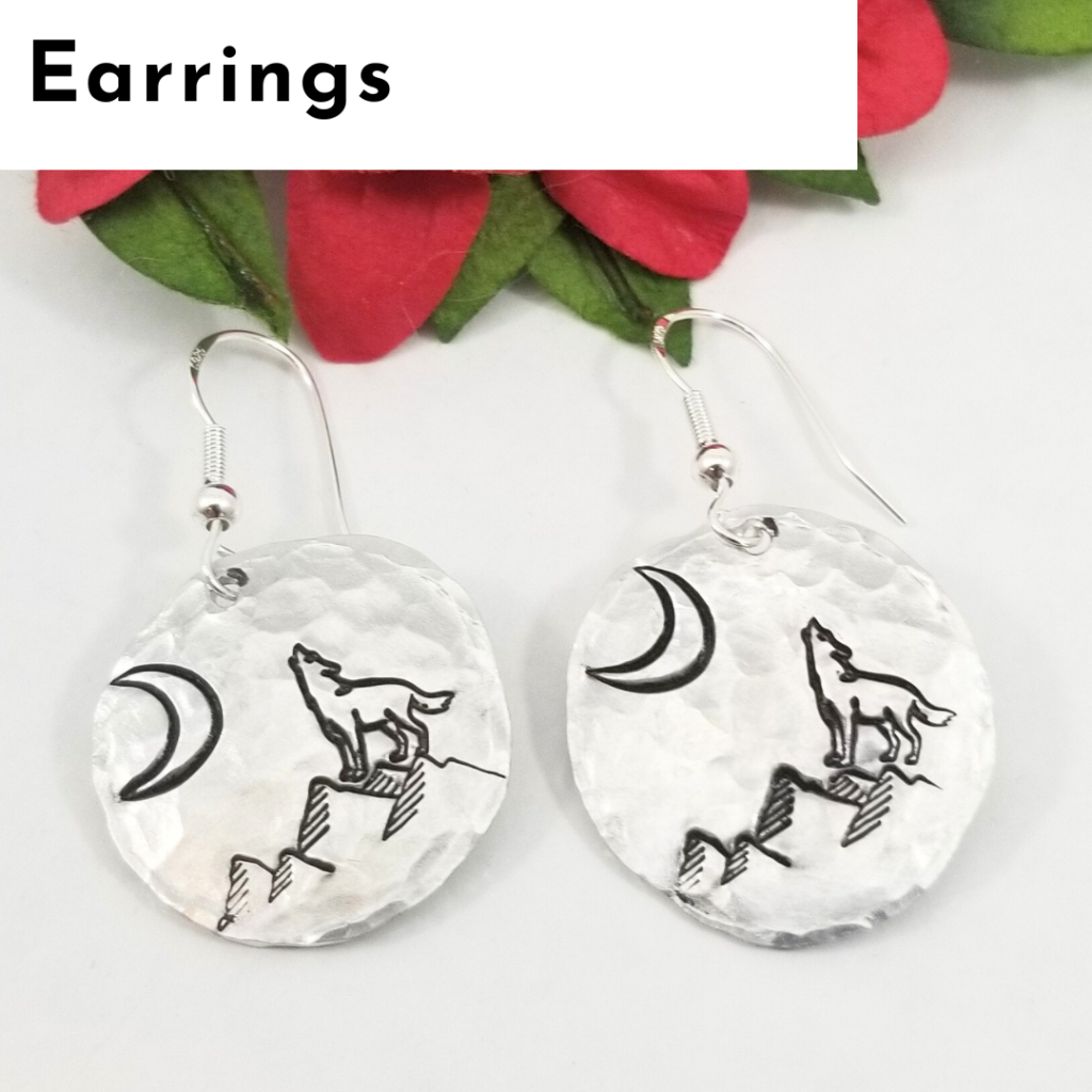 A pair of round, silver, hammered-metal earrings featuring engraved designs of a howling wolf on a mountain with a crescent moon in the sky. These unique earrings offer a touch of personalized jewelry style. Red and green artificial flowers are partially visible behind. Read more at Stamping Cat Studio (stampingcatstudio.com).