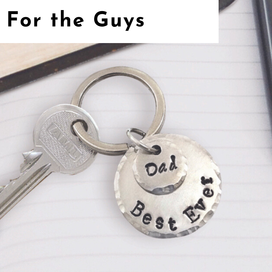 A metal keyring with a key and a round hammered metal tag engraved with “Dad” and “Best Ever” rests on a lined notebook, offering a thoughtful touch of personalized jewelry. The top of the image has text reading “For the Guys.”. Read more at Stamping Cat Studio (stampingcatstudio.com).