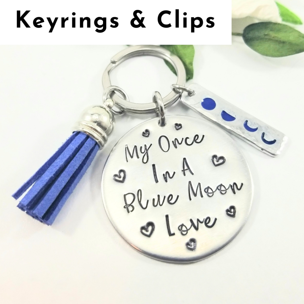 A silver keyring features a round charm stamped with “My Once In A Blue Moon Love” and small heart accents, a blue tassel, and a rectangular charm with blue moon phases—perfect for fans of personalized jewelry. Text above reads, “Keyrings & Clips.”. Read more at Stamping Cat Studio (stampingcatstudio.com).