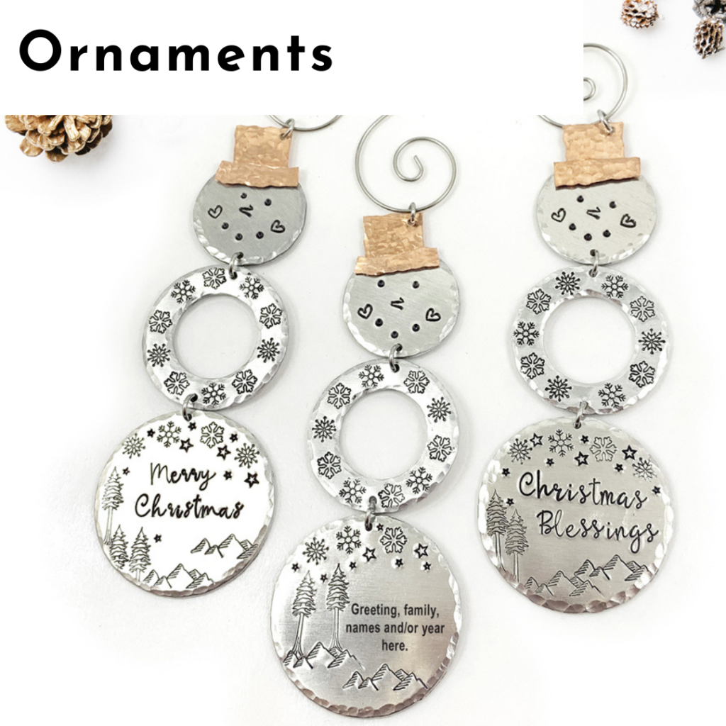 Three snowman-shaped metal ornaments with copper hats and engraved details evoke the charm of personalized jewelry. Two read “Merry Christmas” and “Christmas Blessings”; the third awaits your message. Snowflakes, mountains, and stars accent a white background with pinecones. Read more at Stamping Cat Studio (stampingcatstudio.com).