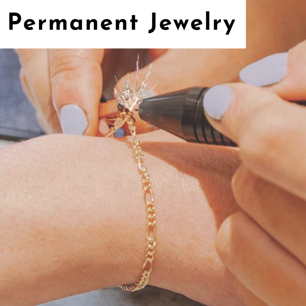 A close-up of hands fusing a gold chain bracelet onto a wrist with a welding tool, creating a small spark. The person's fingernails are painted light blue. Text at the top reads “Permanent Jewelry” in bold black letters, highlighting personalized jewelry. Read more at Stamping Cat Studio (stampingcatstudio.com).
