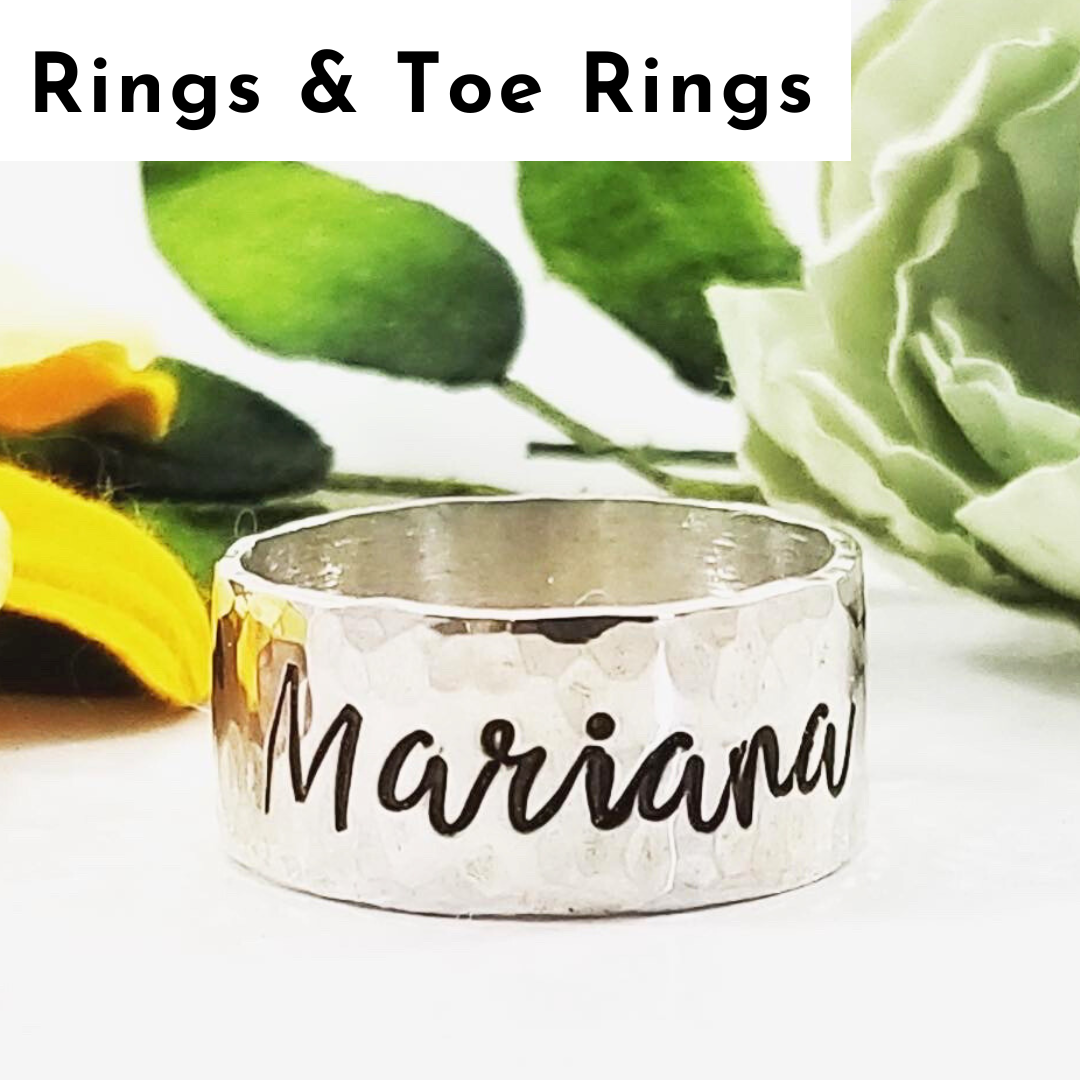 A personalized jewelry piece, this silver ring features "Mariana" engraved in black cursive. Set before green leaves and pale yellow-green flowers, it sits beneath bold black text: "Rings & Toe Rings" on a white background. Read more at Stamping Cat Studio (stampingcatstudio.com).