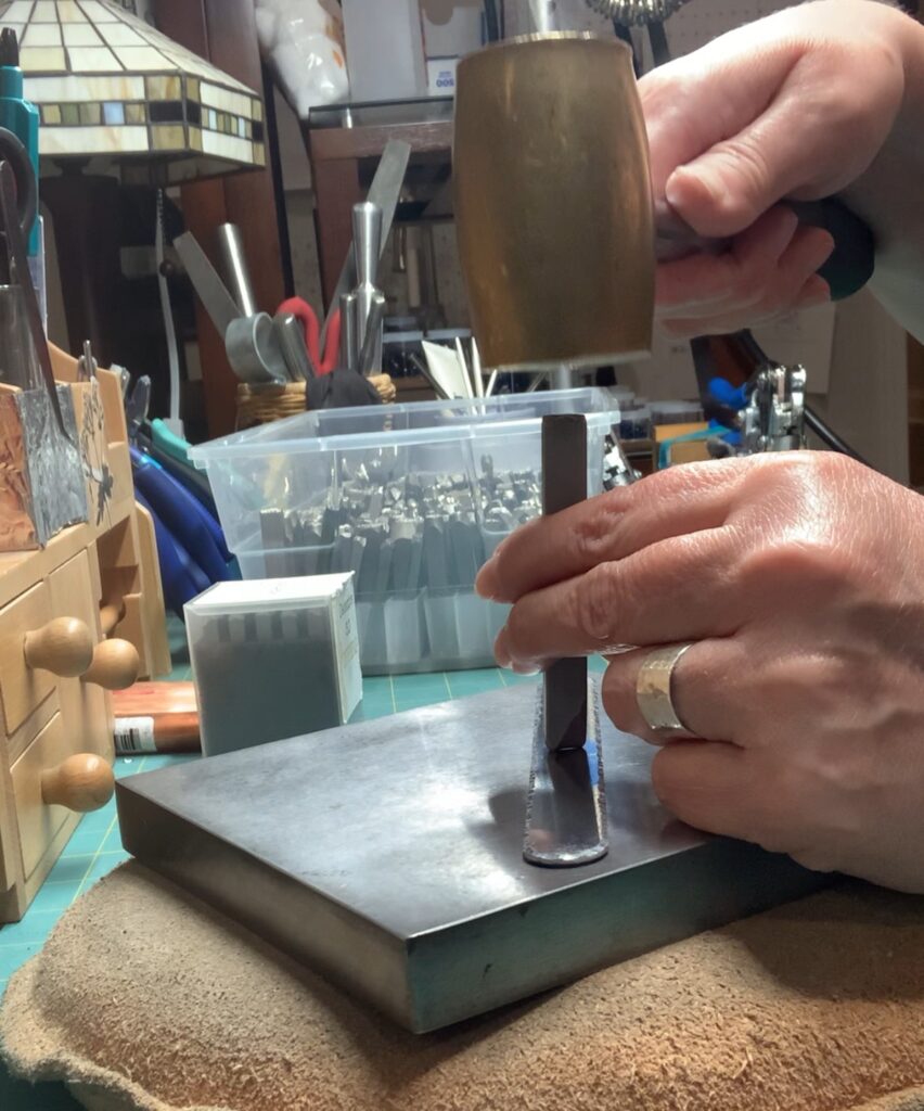 A person uses a brass hammer to strike a metal stamping tool held upright on a metal sheet, making jewelry. The workspace has tools, a wooden drawer, and storage containers in the background. The person wears a silver ring on their left hand. Read more at Stamping Cat Studio (stampingcatstudio.com).