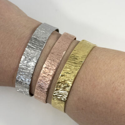 A close-up of a wrist adorned with the Stunning Textured Cuff Bracelet in silver, rose gold, and gold. Each hammered cuff features an uneven surface, aligned side by side against a plain, light background. Read more at Stamping Cat Studio (stampingcatstudio.com).