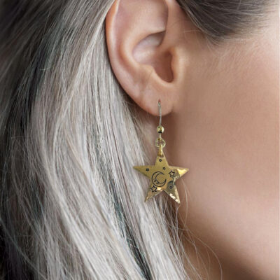A close-up of a person’s ear with long, straight silver hair wearing the Bright Stars And Moon ear nugold, a gold star-shaped earring engraved with a crescent moon and small stars. A softly blurred background highlights its celestial design. Read more at Stamping Cat Studio (stampingcatstudio.com).