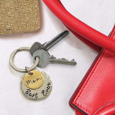 The Best Mom Ever Key Ring, featuring two keys and a round, two-tone charm engraved with "Mom Best Ever," is placed beside a red textured handbag and a gold glittery accessory on a white surface. Read more at Stamping Cat Studio (stampingcatstudio.com).