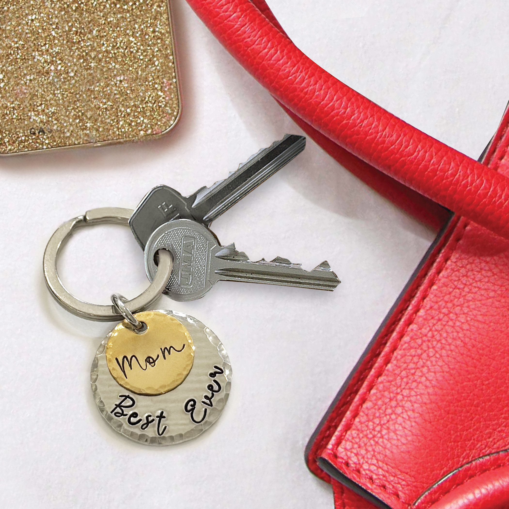 The Best Mom Ever Key Ring, featuring two keys and a round, two-tone charm engraved with "Mom Best Ever," is placed beside a red textured handbag and a gold glittery accessory on a white surface. Read more at Stamping Cat Studio (stampingcatstudio.com).