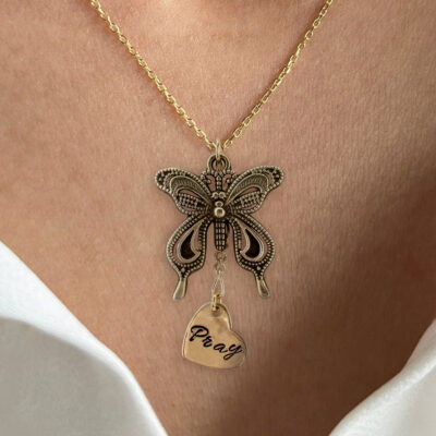 A person in a white off-shoulder top wears the Bronze Butterfly Neck With Pray Heart Charm, featuring a detailed bronze butterfly pendant and a heart charm engraved with "Pray. Read more at Stamping Cat Studio (stampingcatstudio.com).