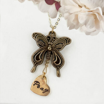 The Bronze Butterfly Neck With Pray Heart Charm main showcases a detailed butterfly pendant alongside a "Pray"-engraved heart charm, displayed against soft white and pale pink artificial flower accents. Read more at Stamping Cat Studio (stampingcatstudio.com).
