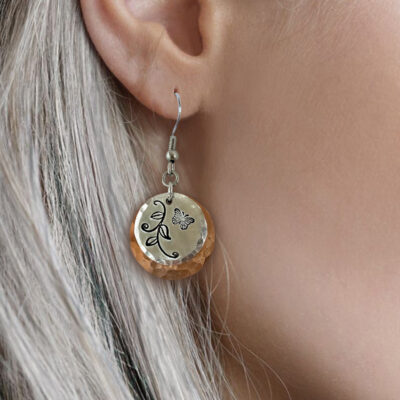 A close-up of a person with long, light gray hair and light skin wearing the Butterfly Garden mixed metal ear—a round, hammered gold-toned earring engraved with a black butterfly and leafy vine design. Read more at Stamping Cat Studio (stampingcatstudio.com).