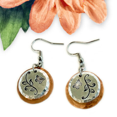 The Butterfly Garden mixed metal earrings feature round, hammered copper and silver discs. Each silver disc is engraved with butterfly and vine designs. These earrings have fishhook wires and are shown below a peach flower with green leaves. Read more at Stamping Cat Studio (stampingcatstudio.com).