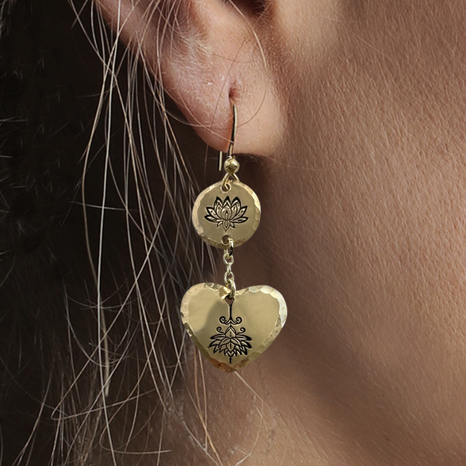 Close-up of a person’s ear wearing Double Lotus Earrings NL—gold dangle earrings with hammered texture, featuring a small disc above a larger heart, both engraved with lotus flowers. Hook fastening; some hair strands visible. Read more at Stamping Cat Studio (stampingcatstudio.com).