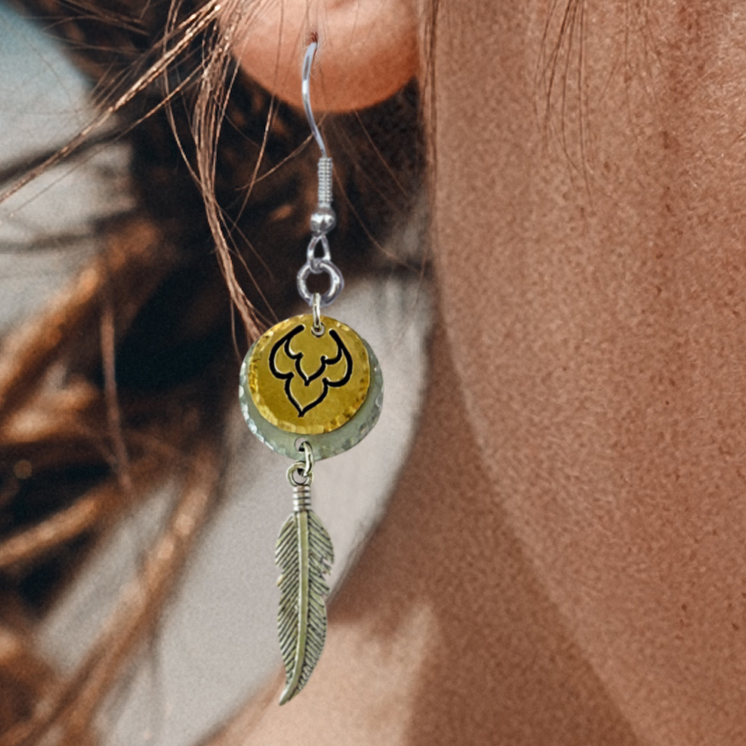 Close-up of a person wearing Free Spirit Feather earrings NL—featuring a yellow circular bead with a black abstract design and a silver feather charm. Brown hair is tied back; background is blurred. Read more at Stamping Cat Studio (stampingcatstudio.com).