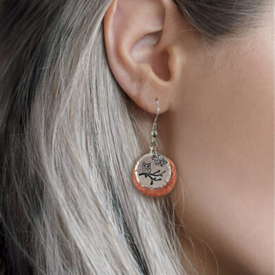 Close-up of a woman with long silver hair wearing the Owl On Branch MM ear: round, hammered copper and silver earrings featuring an engraved owl on a branch and a small wire owl charm above the disk, set against a softly blurred background. Read more at Stamping Cat Studio (stampingcatstudio.com).