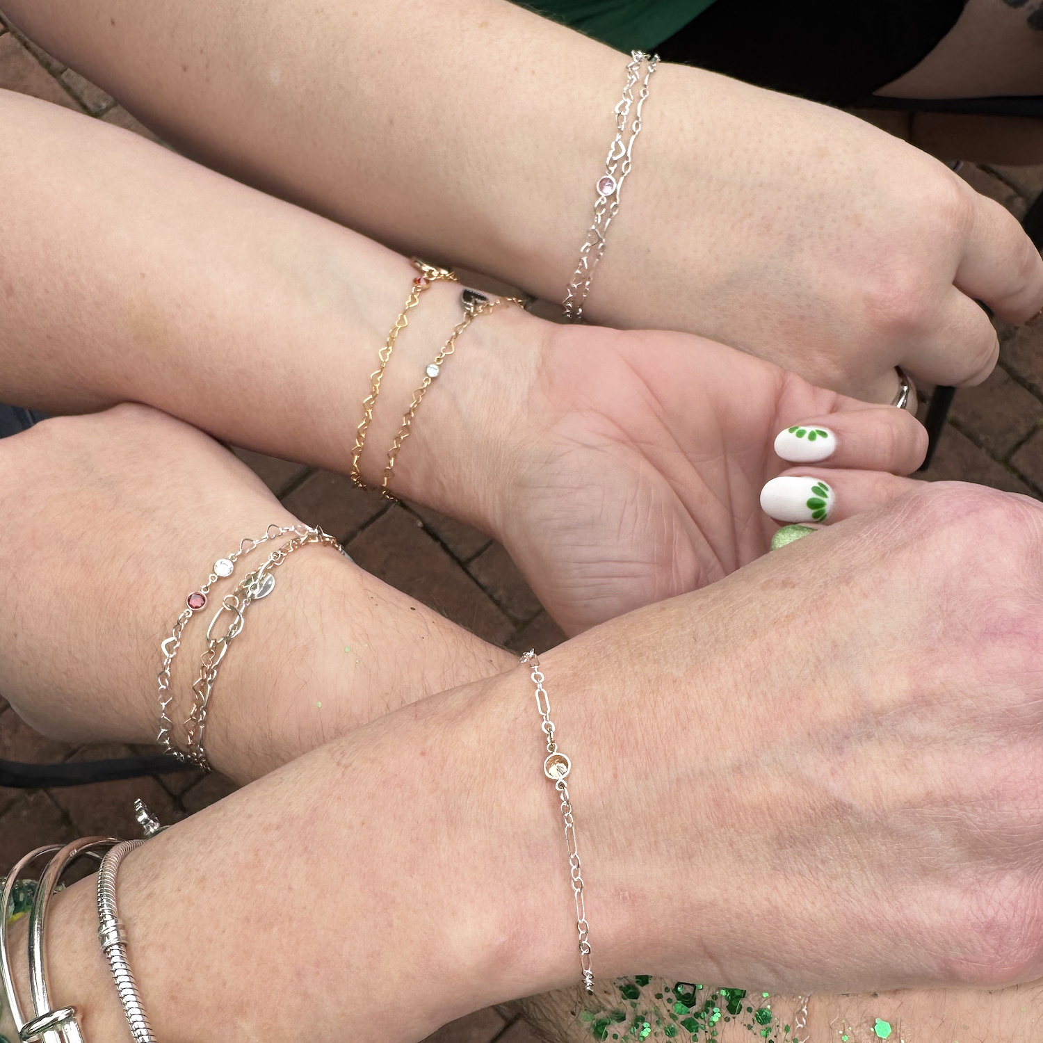 Four forearms adorned with Perm client GFs 1b NL delicate chain bracelets form a circle. The hands display light skin tones; one has white nails with green dots, another features green glitter on the wrist. The background is brick pavement. Read more at Stamping Cat Studio (stampingcatstudio.com).