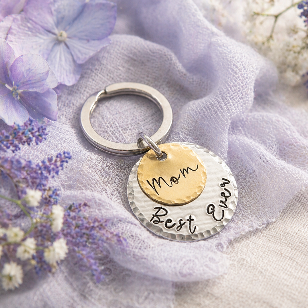 The May 1 Keyring features two layered round metal discs on a silver ring: a gold disc engraved with “Mom” atop a silver disc reading “Best Ever.” Displayed on lavender fabric and accented by purple and white flowers. Read more at Stamping Cat Studio (stampingcatstudio.com).