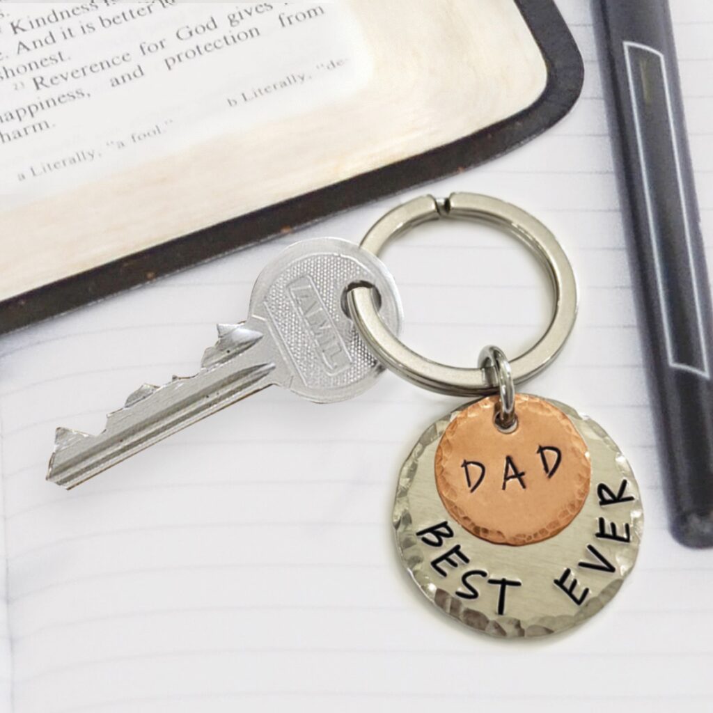 The Best Ever Dad key NL features a silver key on a keyring with two round tags: a copper one engraved "DAD" and a larger silver one reading "BEST EVER," displayed on a lined notebook beside a pen and an open book. Read more at Stamping Cat Studio (stampingcatstudio.com).