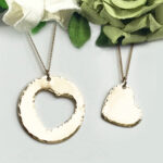 Couples Necklace main: Set of two gold necklaces—one with a large circle pendant and heart cutout, the other with a small heart pendant. Both have textured finishes and are displayed on white with green and white fabric flowers. Read more at Stamping Cat Studio (stampingcatstudio.com).