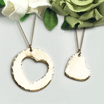 Couples Necklace main: Set of two gold necklaces—one with a large circle pendant and heart cutout, the other with a small heart pendant. Both have textured finishes and are displayed on white with green and white fabric flowers. Read more at Stamping Cat Studio (stampingcatstudio.com).