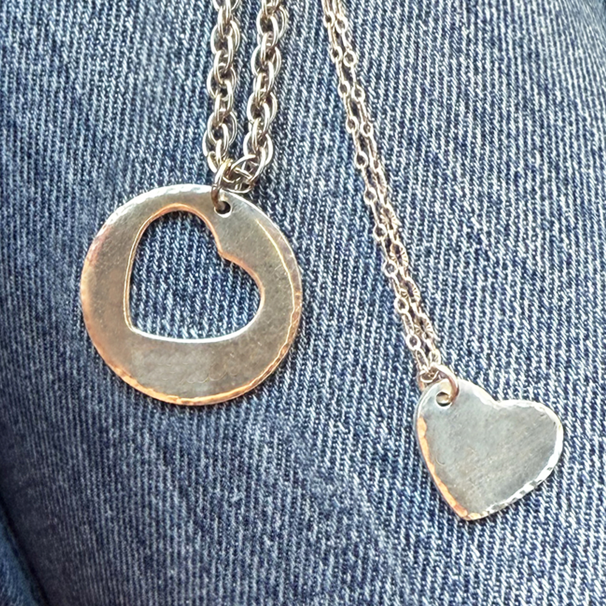 Two silver Couples Necklace pendants rest on blue denim: one round with a heart cutout, the other a small heart charm, both with a rustic hammered texture—perfect his and hers gift for boyfriend and girlfriend. Read more at Stamping Cat Studio (stampingcatstudio.com).