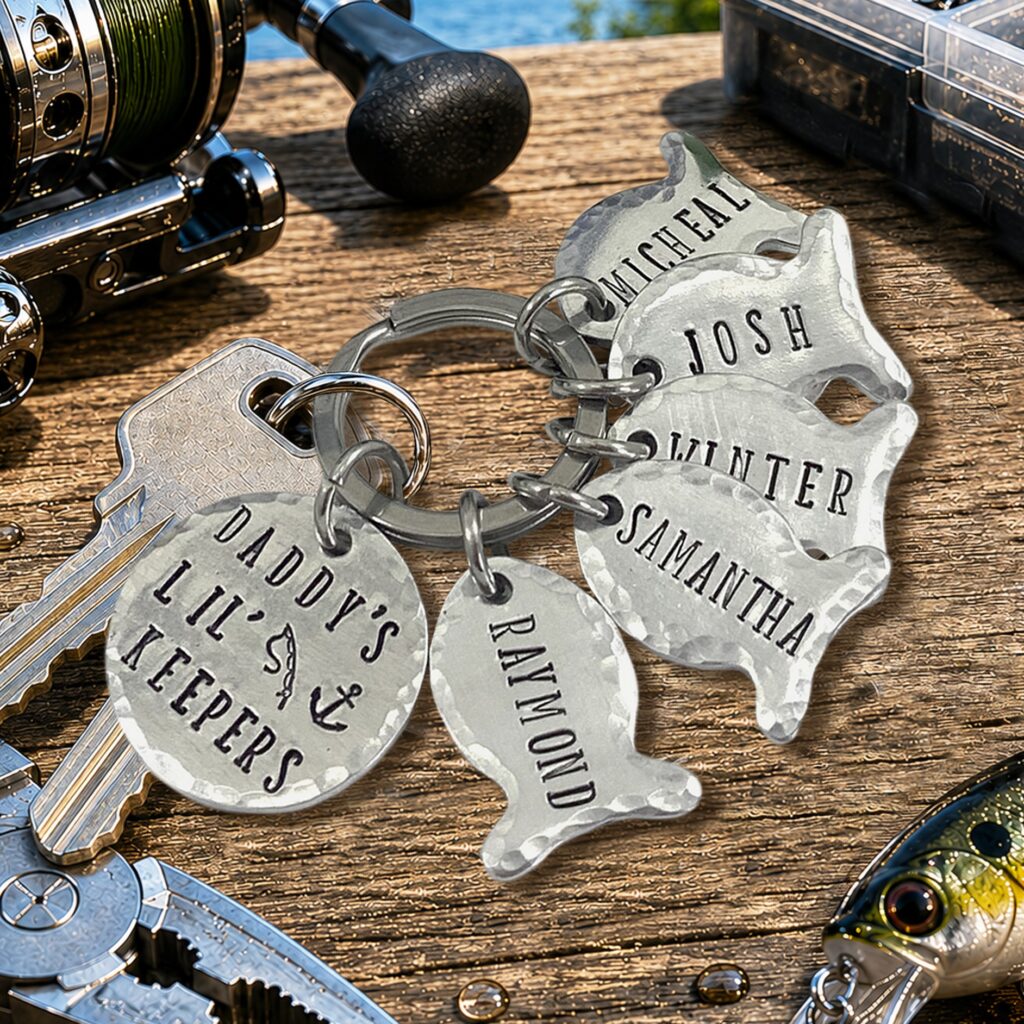 The Daddys NLjpg silver, fish-shaped keychains—engraved with custom names or messages—hang on a key ring with house keys, displayed on a wooden table next to fishing gear and a scenic water backdrop. Read more at Stamping Cat Studio (stampingcatstudio.com).
