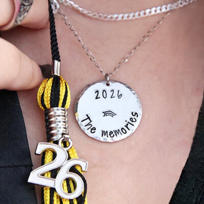 A close-up of the Pers Graduation neck or key, showcasing a silver round pendant engraved “2026 The memories,” a small graduation cap, black and yellow tassel, and a metallic “26” charm—a perfect personalized graduation gift. Read more at Stamping Cat Studio (stampingcatstudio.com).