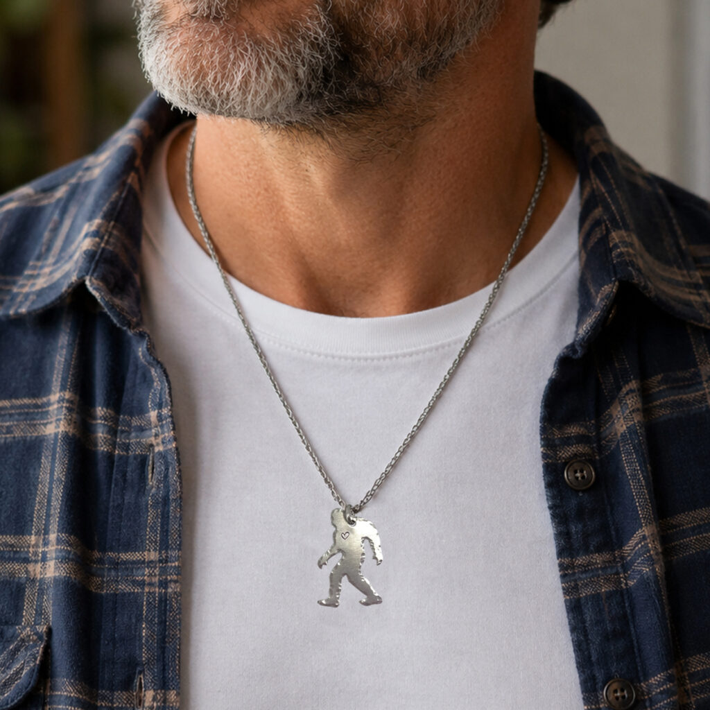 A man with a gray beard wears a white T-shirt, blue plaid shirt, and the "Whimsical bigfoot lifestyle" silver necklace featuring a Bigfoot-shaped pendant. The image shows only his neck and upper chest. Read more at Stamping Cat Studio (stampingcatstudio.com).