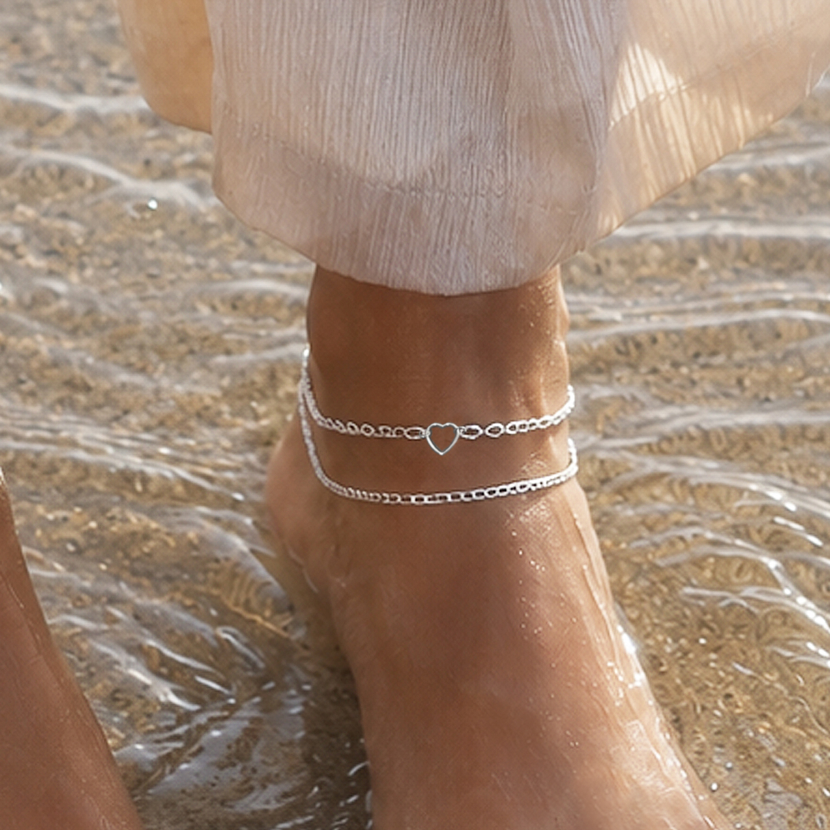 A close-up of a bare ankle adorned with the Perm ankle July NL rev double-layered silver anklet with a heart charm, standing in shallow beach water. Sheer fabric drapes part of the ankle as sunlight shimmers on the wet sand. Read more at Stamping Cat Studio (stampingcatstudio.com).