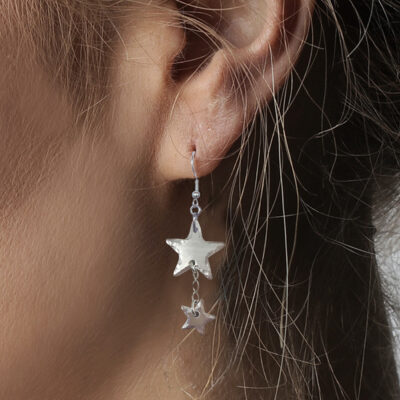 A close-up showcases a person’s ear adorned with the Shining Star NL—a dangling silver earring featuring two metallic star charms, with a larger top star and a smaller star below, accented by light brown hair strands. Read more at Stamping Cat Studio (stampingcatstudio.com).