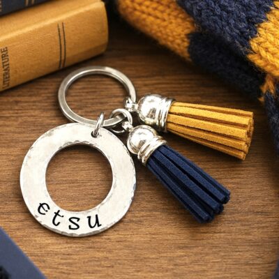 The NL Tassel key ring, featuring a hammered silver ring stamped "etsu" and two suede tassels in navy blue and yellow, rests on a knitted scarf beside a stack of books. Read more at Stamping Cat Studio (stampingcatstudio.com).