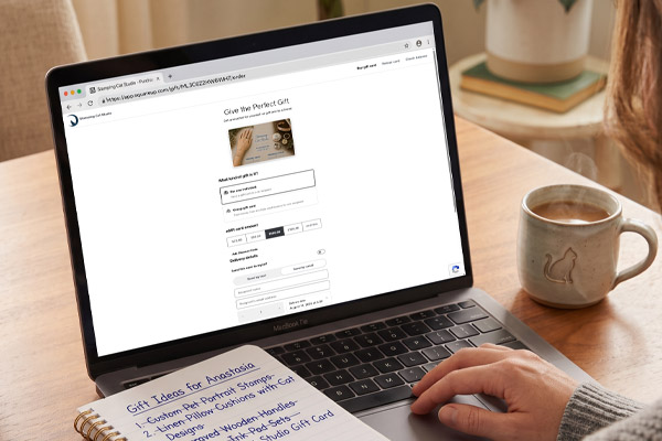 A person uses a laptop to browse the Stamping Cat Studio online website to purchase a gift card. Beside the MacBook, a handwritten note titled Gift Ideas for Anastasia lists six suggestions. A cat-themed mug and a pen rest on the wooden table, creating a cozy and inviting atmosphere. Read more at Stamping Cat Studio (stampingcatstudio.com).