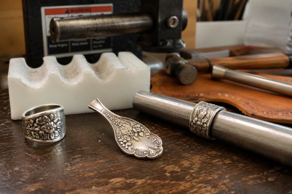 A vintage flatware spoon ring from Stamping Cat Studio rests on a wooden table alongside a pendant, another ring on a mandrel, a white tool holder, metal tools, and leatherwork—evoking the creative atmosphere of a jewelry crafting workspace, and helping to answer the question what is a spoon ring. Read more at Stamping Cat Studio (stampingcatstudio.com).