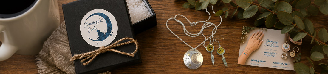A black jewelry box labeled “Stamping Cat Studio” sits by a mug, fabric, silver necklace, two pairs of earrings, leaves, and a stamping cat studio gift card for personalized and permanent jewelry on a wooden table. The gift card features an illustrated hand adorned with stacked rings. Read more at Stamping Cat Studio (stampingcatstudio.com).
