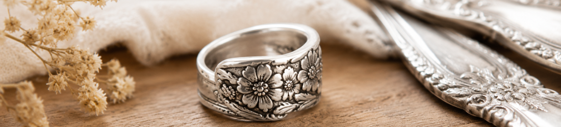 A vintage flatware jewelry spoon ring from Stamping Cat Studio, featuring intricate, raised floral details, sits on a wooden surface beside ornate silverware, dried flowers, and a cream-colored cloth. Read more at Stamping Cat Studio (stampingcatstudio.com).