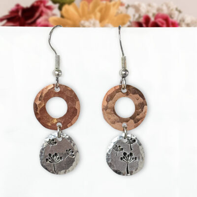 A pair of Dandelion Dream Dangle Earrings featuring hammered copper circles accented with delicate stamped botanical designs on top, linked to hammered silver ovals below with matching botanical motifs. A soft-focus bouquet of pink, yellow, and white flowers provides a colorful background. Read more at Stamping Cat Studio (stampingcatstudio.com).