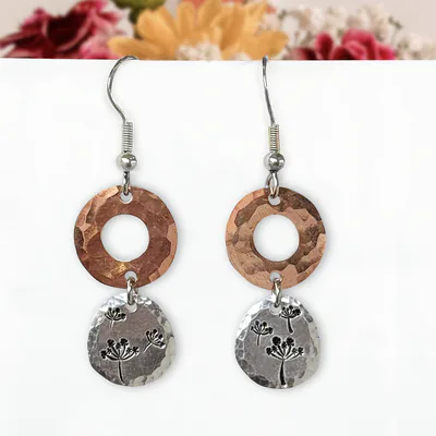 A pair of Dandelion Dream Dangle Earrings featuring hammered copper circles accented with delicate stamped botanical designs on top, linked to hammered silver ovals below with matching botanical motifs. A soft-focus bouquet of pink, yellow, and white flowers provides a colorful background. Read more at Stamping Cat Studio (stampingcatstudio.com).