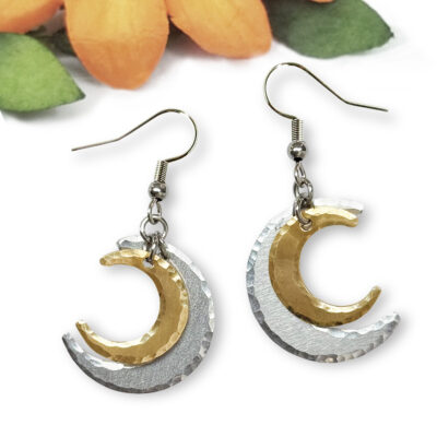 Introducing the Double Crescent Moon Ear Main: a pair of dangling earrings featuring a smaller gold crescent layered over a larger hammered silver crescent, beautifully displayed against a softly blurred background accented with orange and green floral elements. Read more at Stamping Cat Studio (stampingcatstudio.com).