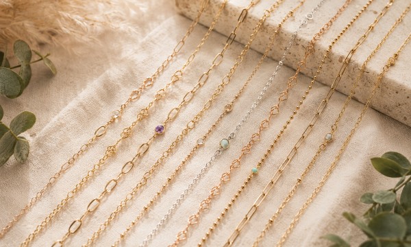 Multiple delicate gold and silver permanent jewelry chains from Stamping Cat Studio, accented with small gemstones and beads, are neatly arranged in parallel lines on a beige fabric. Green eucalyptus leaves decorate the corners, creating a soft, elegant display reminiscent of a boutique Permanent Jewelry Studio. Read more at Stamping Cat Studio (stampingcatstudio.com).