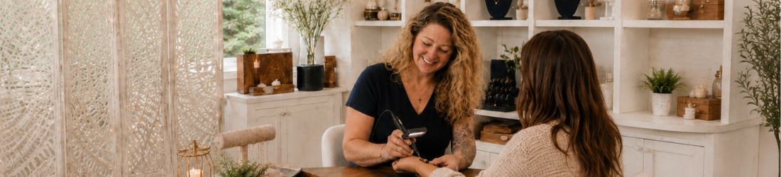 Shannon, a woman with curly blonde hair smiles as she welds a custom-fit permanent bracelet at Stamping Cat Studio in Gray, Tennessee. They sit at a wooden table in a cozy room filled with shelves, plants, candles, and soft natural light. Book your permanent jewelry appointment today at Stamping Cat Studio to experience this inviting atmosphere for yourself!. Read more at Stamping Cat Studio (stampingcatstudio.com).