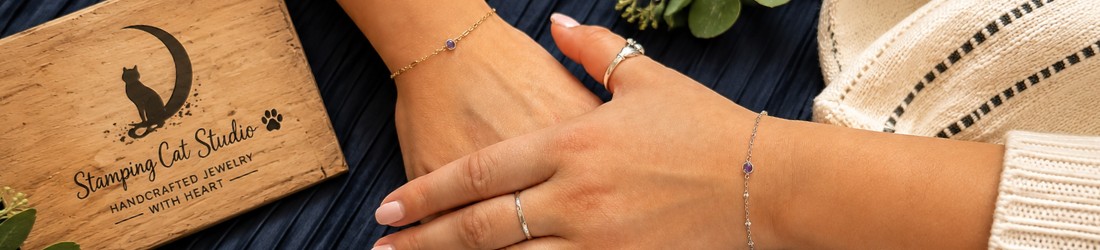 Two hands adorned with delicate permanent bracelets and rings rest on dark fabric, one wearing a cream sweater. To the left sits a wooden box featuring the Stamping Cat Studio logo—a black cat and the words “Handcrafted Jewelry With Heart”—all surrounded by small flowers and greenery. This inviting scene beautifully illustrates what to expect at a permanent jewelry appointment at Stamping Cat Studio, making it an ideal choice for those seeking Permanent Jewelry in Tri-Cities TN or wishing to book an appointment in Johnson City TN. Read more at Stamping Cat Studio (stampingcatstudio.com).