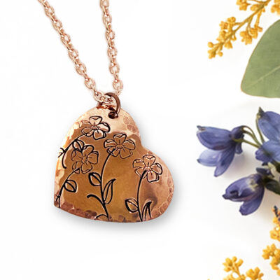 The Heart Bloom Copper Neck main is a hammered copper heart-shaped pendant on a matching link chain, featuring etched line art of three small flowers, displayed on a white background next to sprigs of small blue and yellow flowers. Read more at Stamping Cat Studio (stampingcatstudio.com).