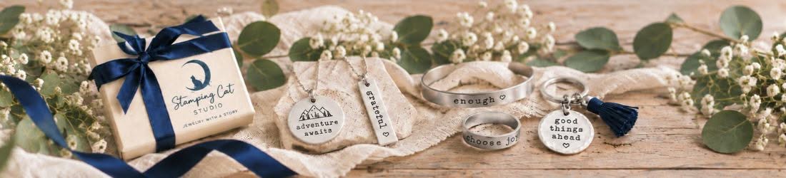 A gift box with a navy blue ribbon sits beside two necklaces, a cuff bracelet, a ring, and a keychain on a white fabric-covered table adorned with green eucalyptus leaves and baby’s breath. Read more at Stamping Cat Studio (stampingcatstudio.com).